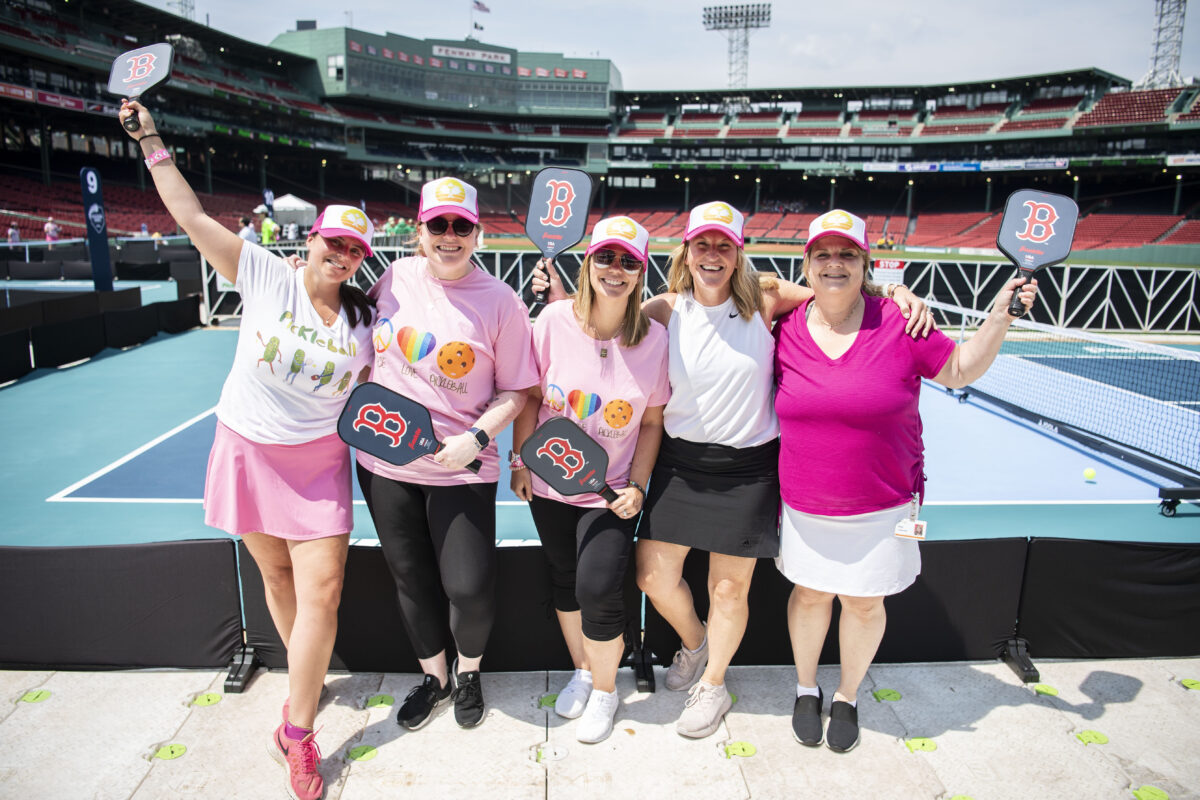 A group of five pickleball players smiling and holding up Boston Red Sox-branded paddles. They are standing in front of a pickleball court inside Fenway Park.